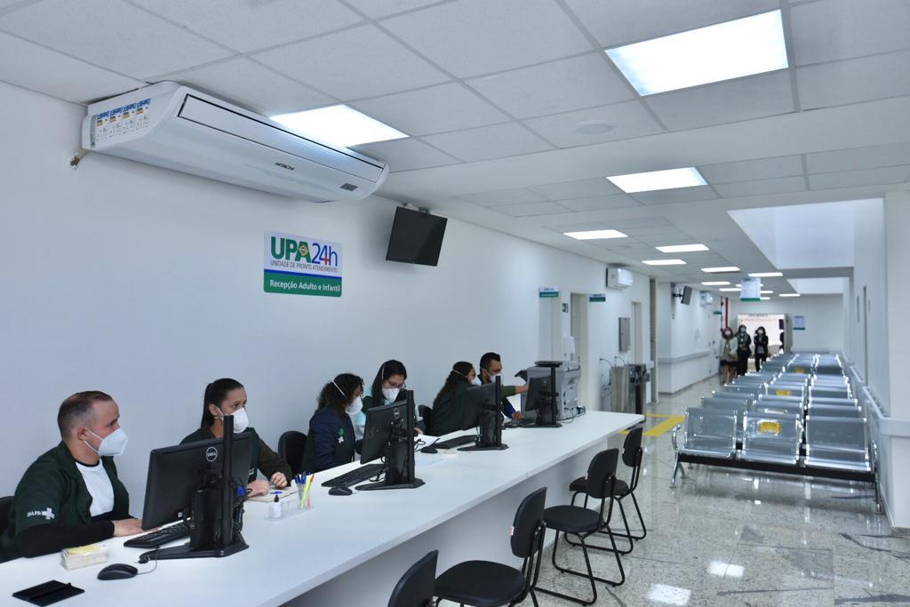 FOTO DE RECEPÇÃO -  Em primeiro plano temos uma longa mesa com recepcionistas  uniformizados em verde escuro, sentados diante do computador, um homem e três mulheres,  ao centro um longo corredor com um homem caminhando e à direita uma série de cadeiras metálicas prateadas